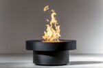 large, black, round, steel gas fire pit known as the Badlands with tall flames in white studio